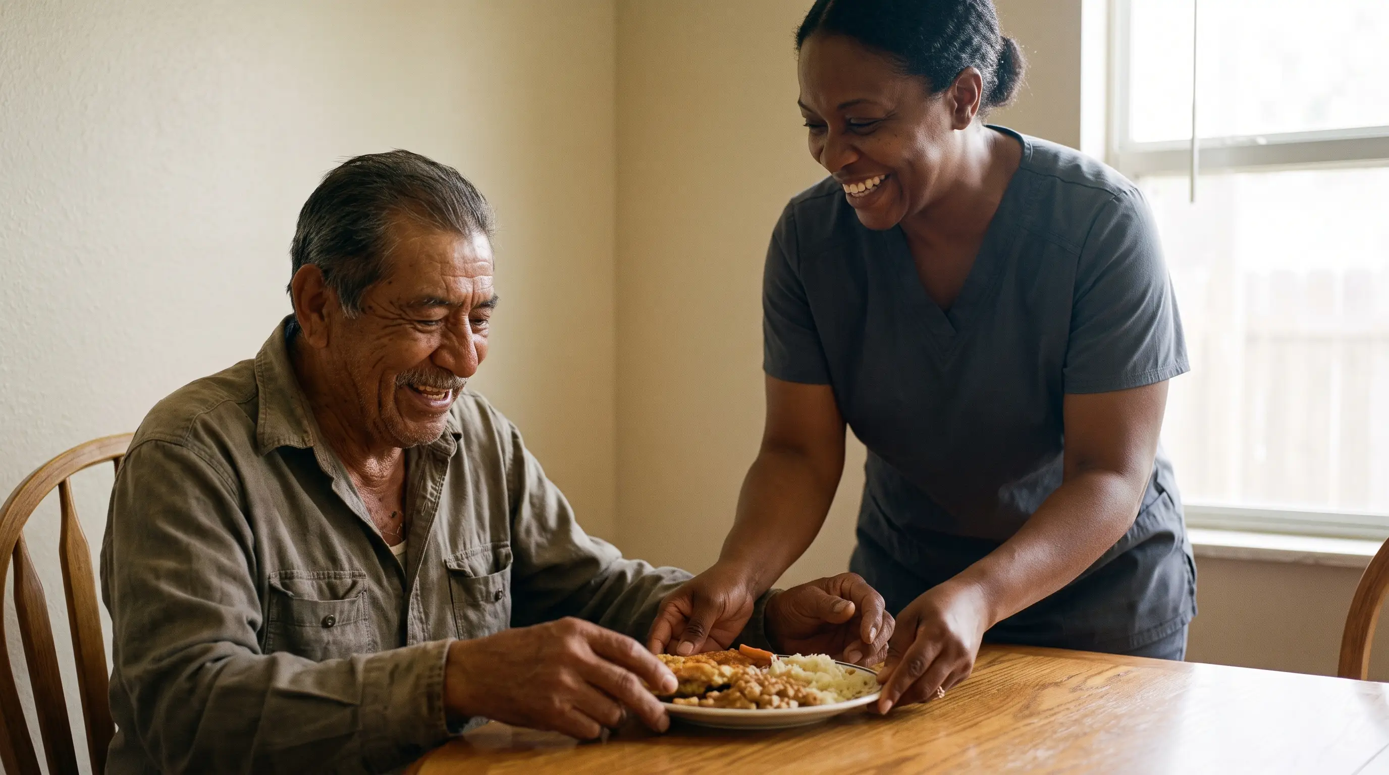 A man in his 80s laughs at his kitchen table as his home care aide sets down his lunch